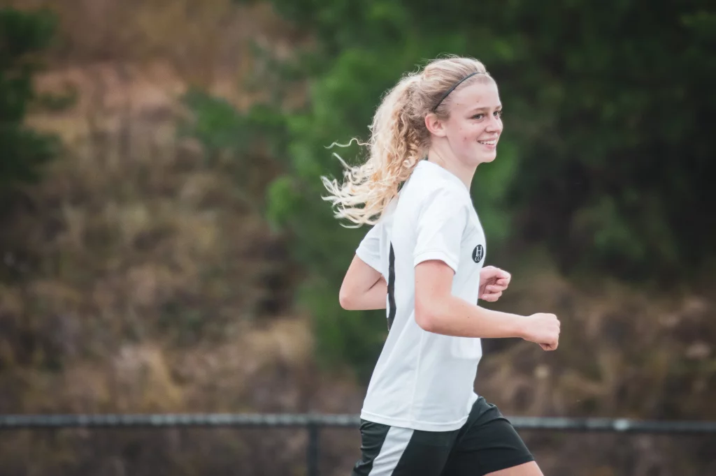 United FC - Furman Region -: Teenage girl with blonde hair in a ponytail, wearing a white soccer jersey and black shorts, running outdoors on a sports field with greenery in the background.