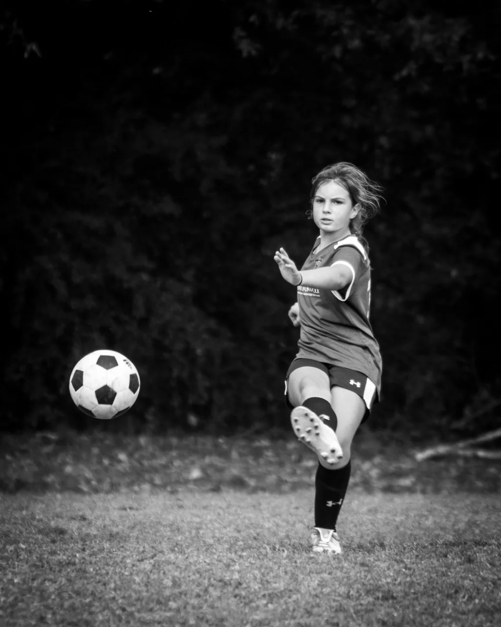 : Black and white photo of a young soccer player kicking a ball on a grassy field, with trees in the background.