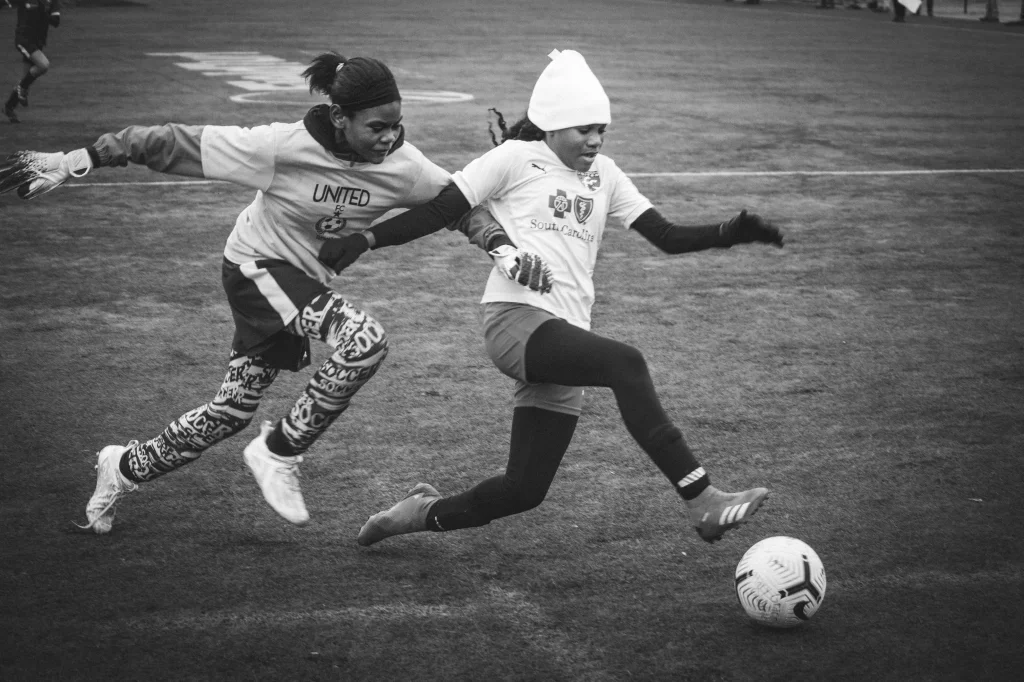 : Two young soccer players in action on a field, one kicking the ball while the other tries to defend, both wearing winter hats and gloves.
