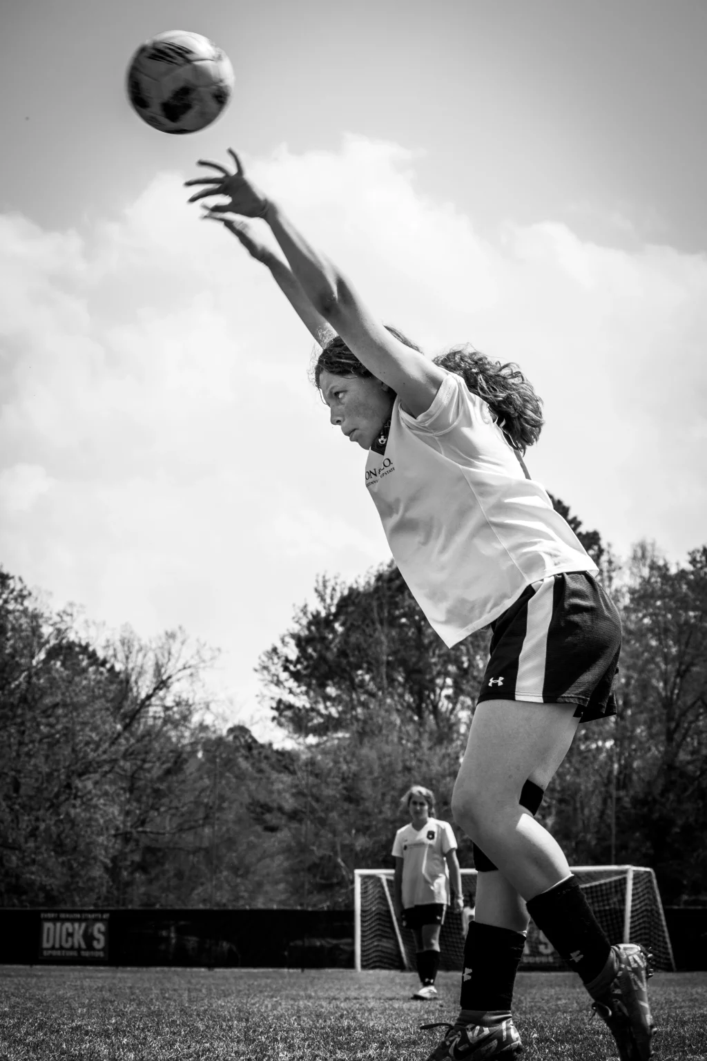 : Black and white photo of a young soccer player in mid-action, throwing a soccer ball overhead during a game, with another player and a goal visible in the background.