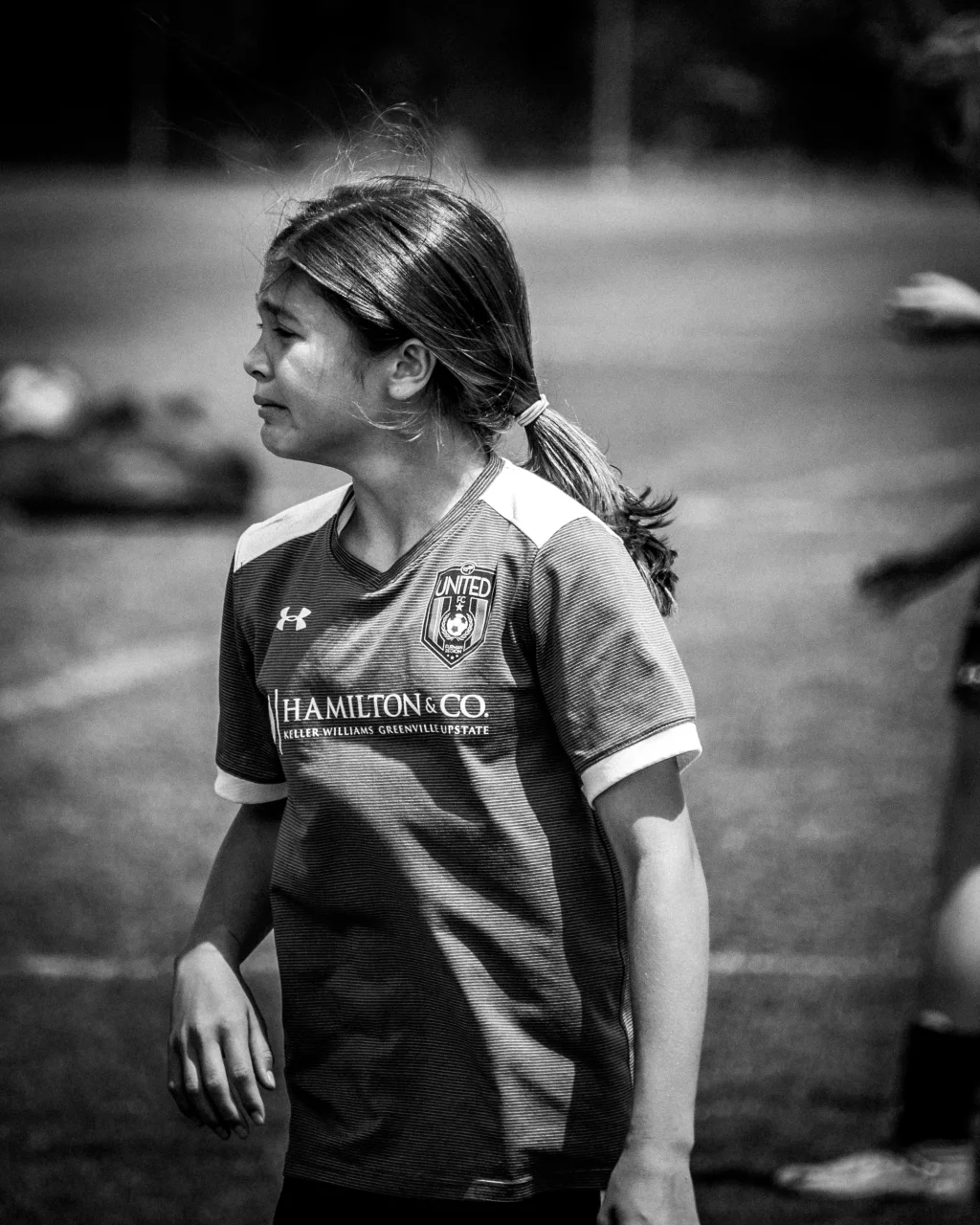 : Black and white photo of a young girl with a ponytail wearing a "United FC" soccer jersey, standing on a soccer field and looking to the left.