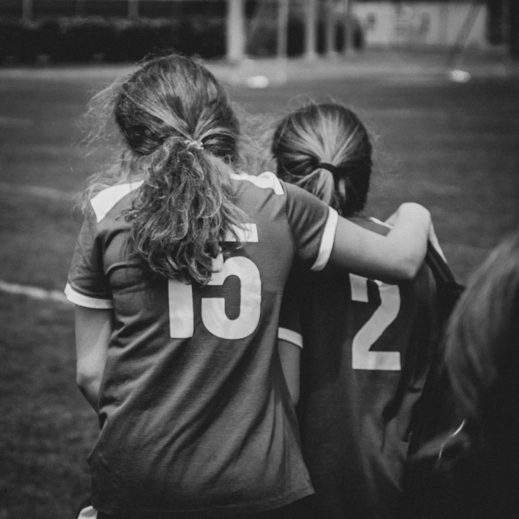 : Black and white photo of two young soccer players, seen from behind, with one arm around the other's shoulder. They wear jerseys with the numbers 15 and 2.