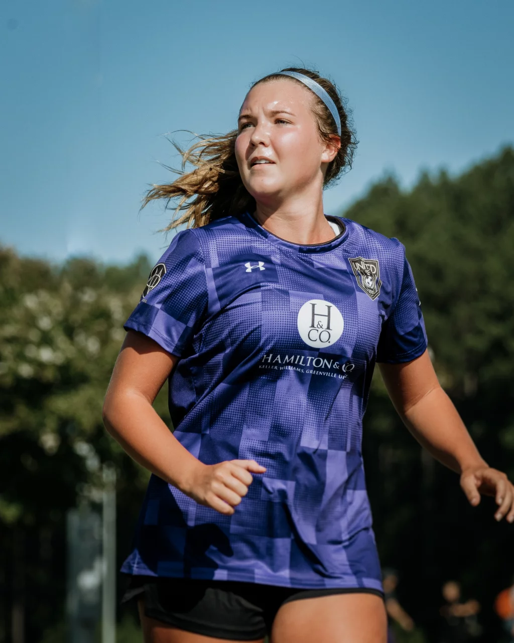 United FC - Furman Region -: Young woman in a purple soccer jersey running outdoors, with trees and blue sky in the background.