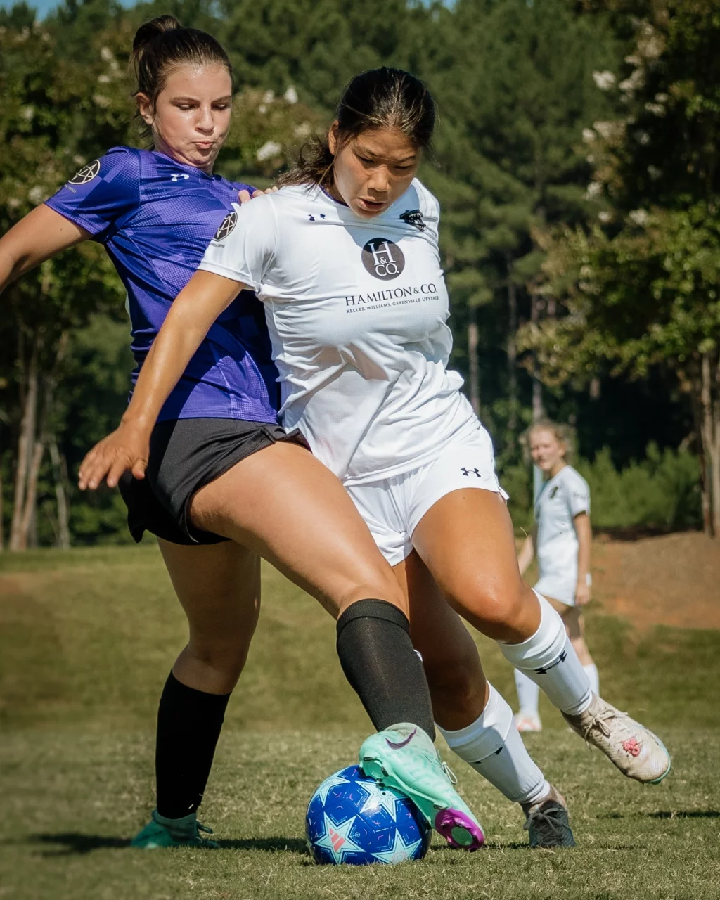United FC - Furman Region -: Two female soccer players compete intensely for the ball on a grassy field, one in a white uniform and the other in purple, with another player in white in the background.