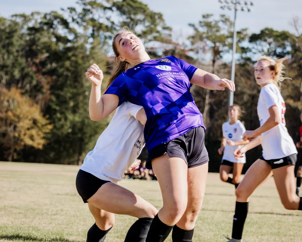 United FC - Furman Region -: Two female soccer players in action, one in a purple jersey and the other in a white jersey, collide while competing for the ball on a grassy field, with other players and trees in the background.