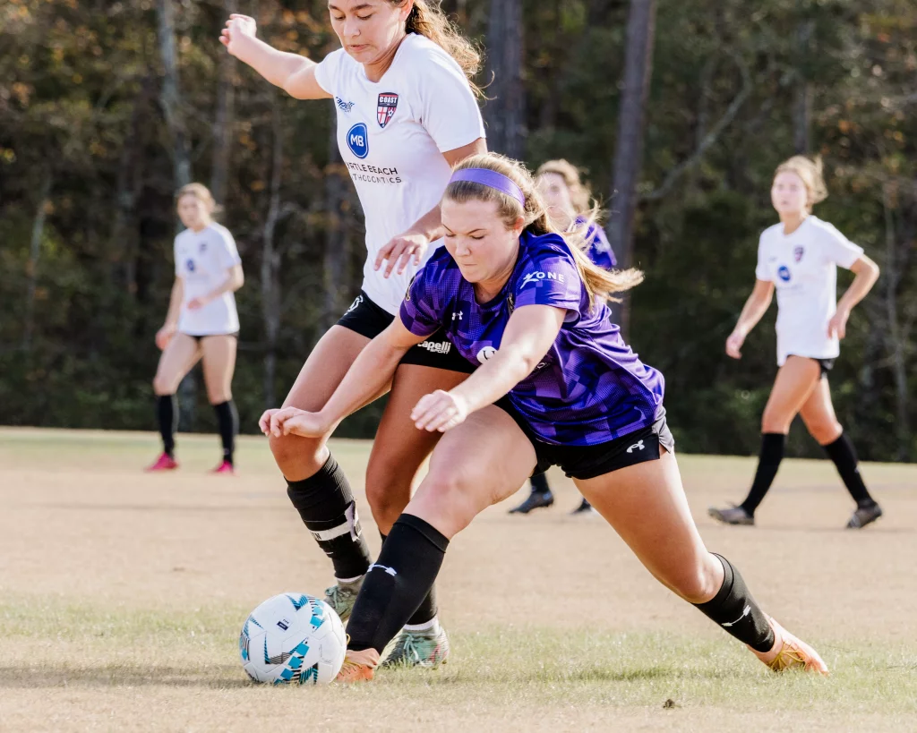 United FC - Furman Region -: Two female soccer players compete for the ball, with one in a purple jersey lunging forward and the other in a white jersey closely defending. Other players and trees are visible in the background.