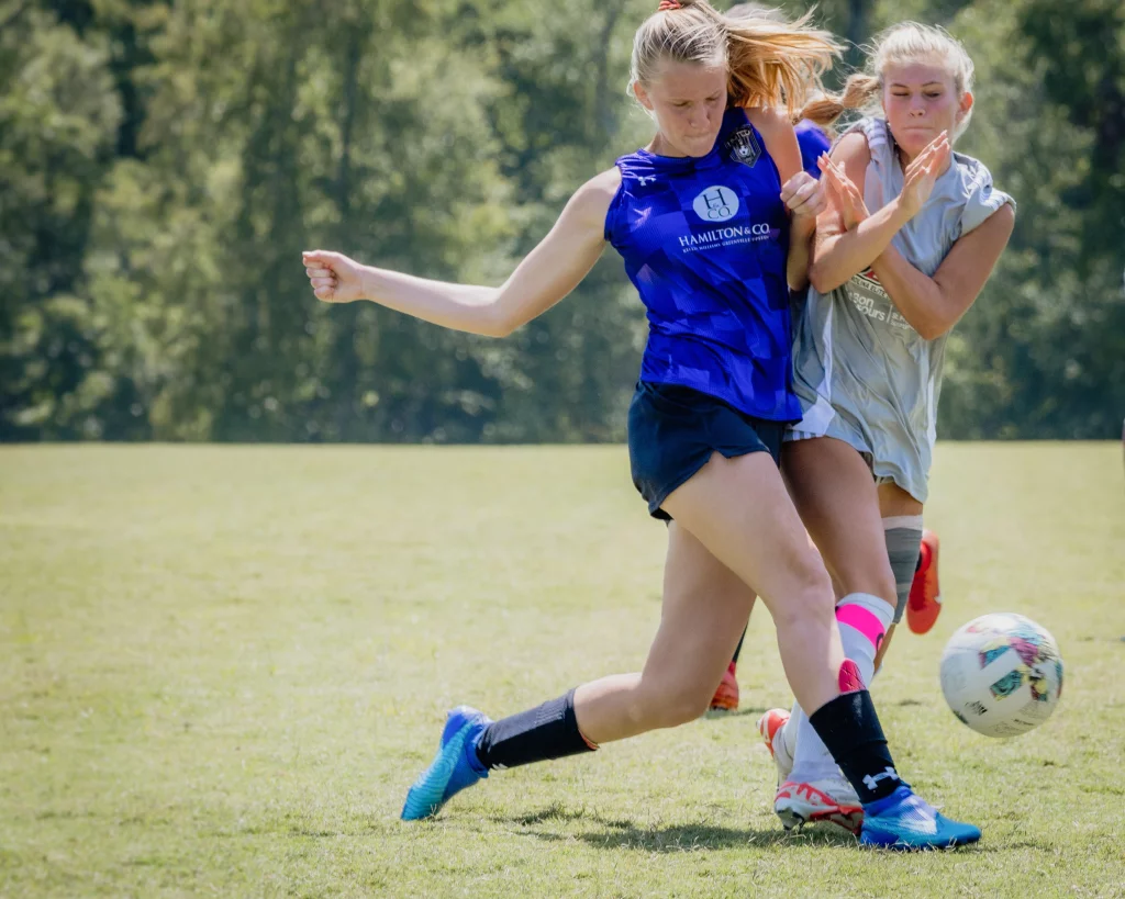 United FC - Furman Region -: Two teenage soccer players compete for the ball on a grassy field, one wearing a blue jersey and the other in gray, with trees in the background.