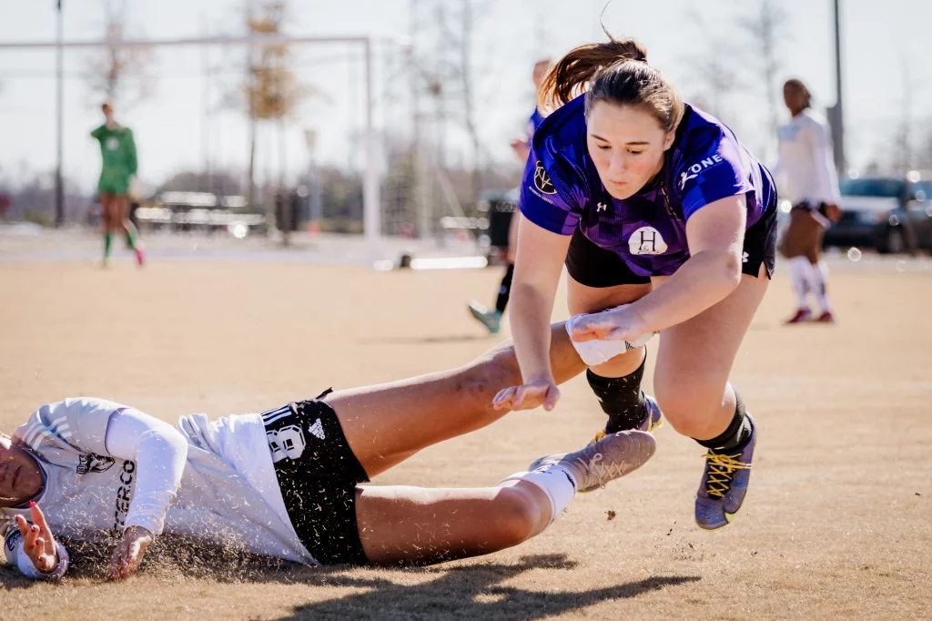United FC - Furman Region -: A soccer player in a purple jersey is mid-air, falling forward, while another player in a white jersey is on the ground after a slide tackle. The action takes place on a dirt field with other players and a goal visible in the background.