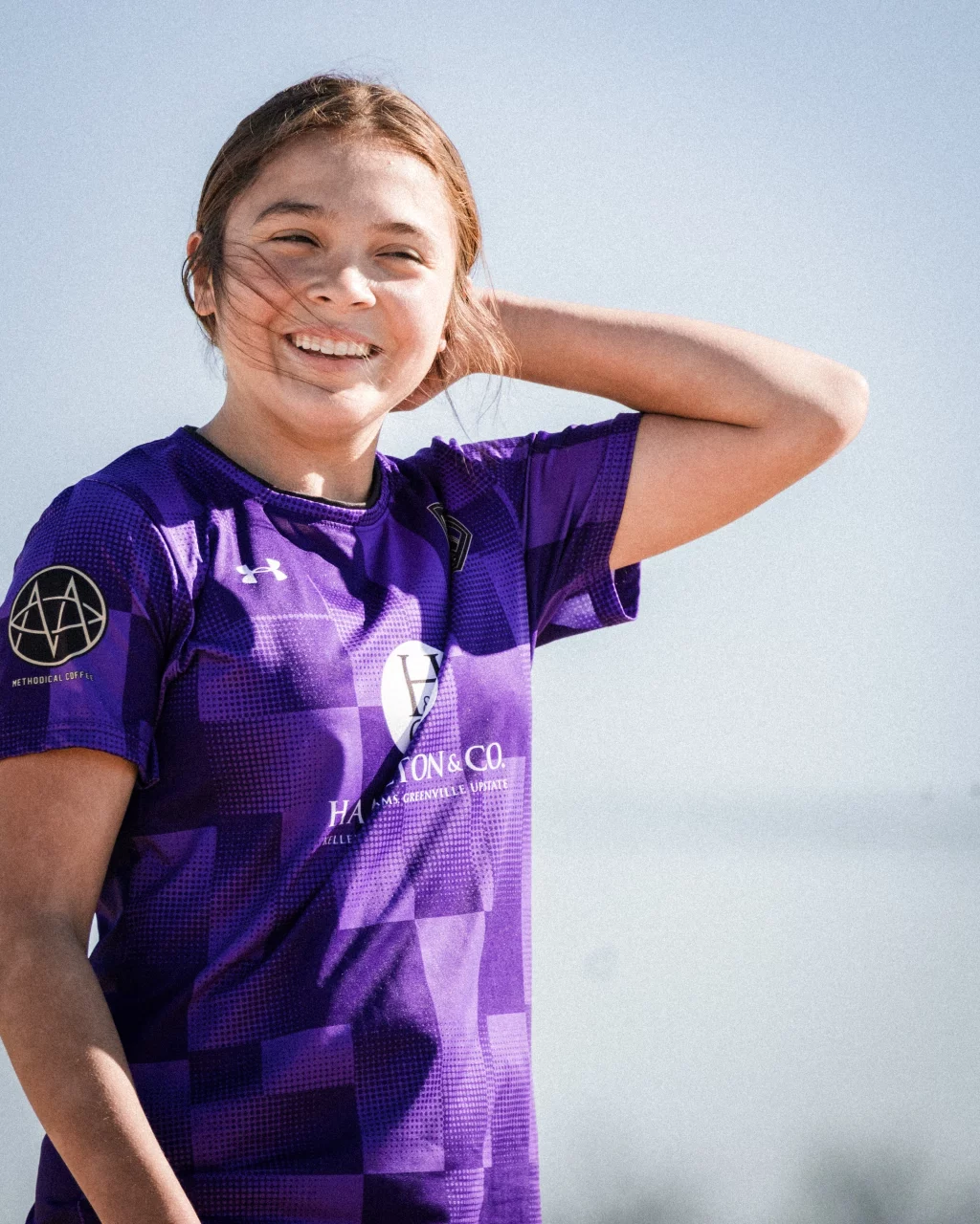 United FC - Furman Region -: A smiling young person wearing a purple sports jersey stands outdoors, with one hand behind their head and wind blowing their hair.
