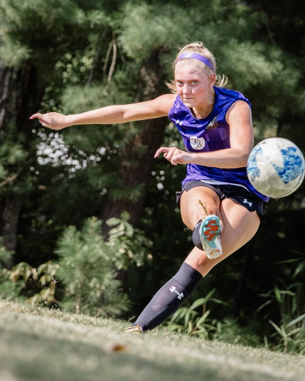 United FC - Furman Region -: A female soccer player in a purple jersey and black shorts is mid-kick, focusing intently on a soccer ball in motion. She is outdoors on a grassy field with trees in the background.