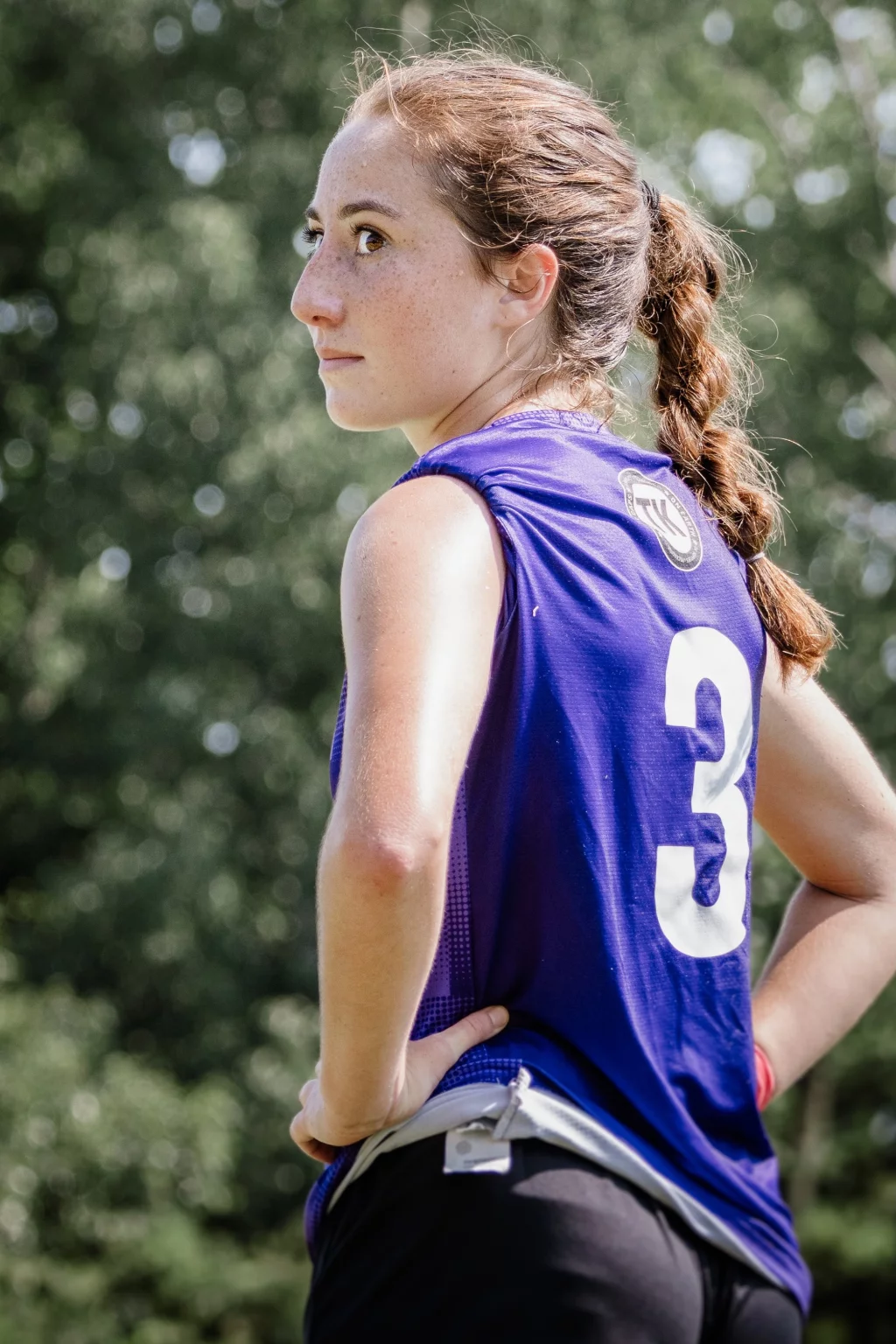United FC - Furman Region -: A young woman with braided hair stands outdoors, wearing a sleeveless purple sports jersey with the number 3 on the back, hands on hips, looking to the side. Trees and greenery are blurred in the background.