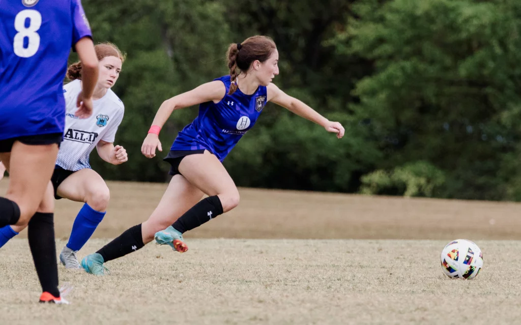 United FC - Furman Region -: A youth soccer player in a blue jersey sprints toward a colorful soccer ball on a grassy field, with other players in blue and white jerseys nearby and trees in the background.