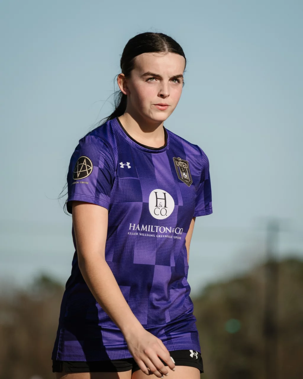 United FC - Furman Region -: A young female soccer player in a purple jersey stands outdoors, looking focused. The sky is clear and blue in the background.