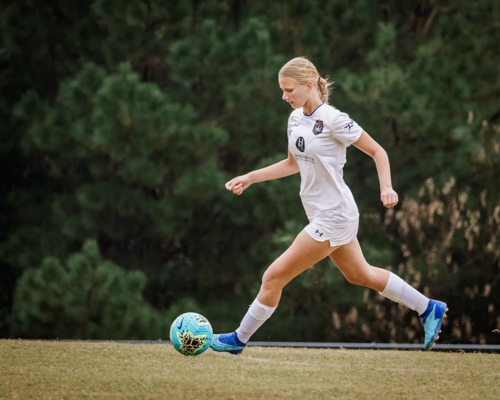 United FC - Furman Region -: A soccer player in a white uniform running on a grassy field, preparing to kick a blue soccer ball, with trees in the background.