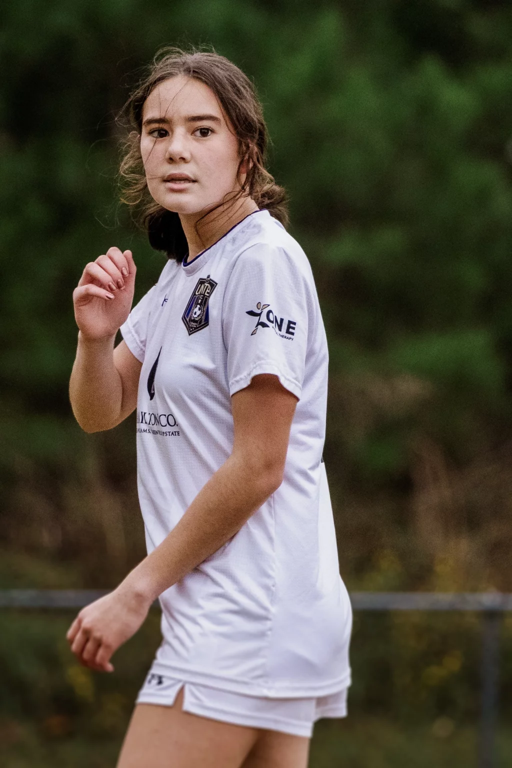 United FC - Furman Region -: A female soccer player wearing a white uniform is standing outdoors on a field, looking to the side with a focused expression. Trees and blurred background are visible behind her.