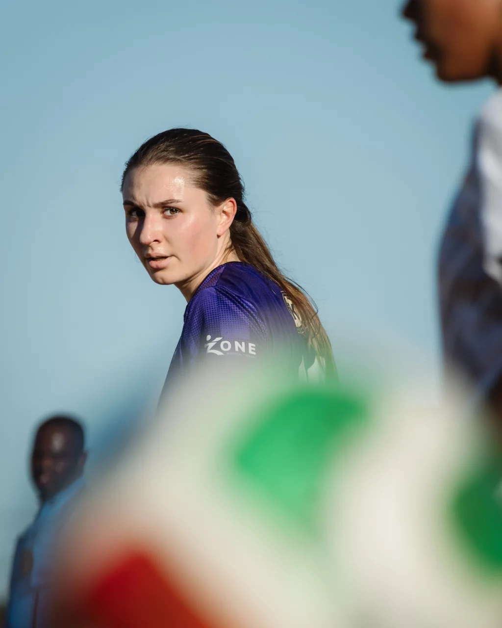 United FC - Furman Region -: A female soccer player in a purple jersey looks over her shoulder with a serious expression, with a blurred foreground and other people partially visible in the background against a clear blue sky.