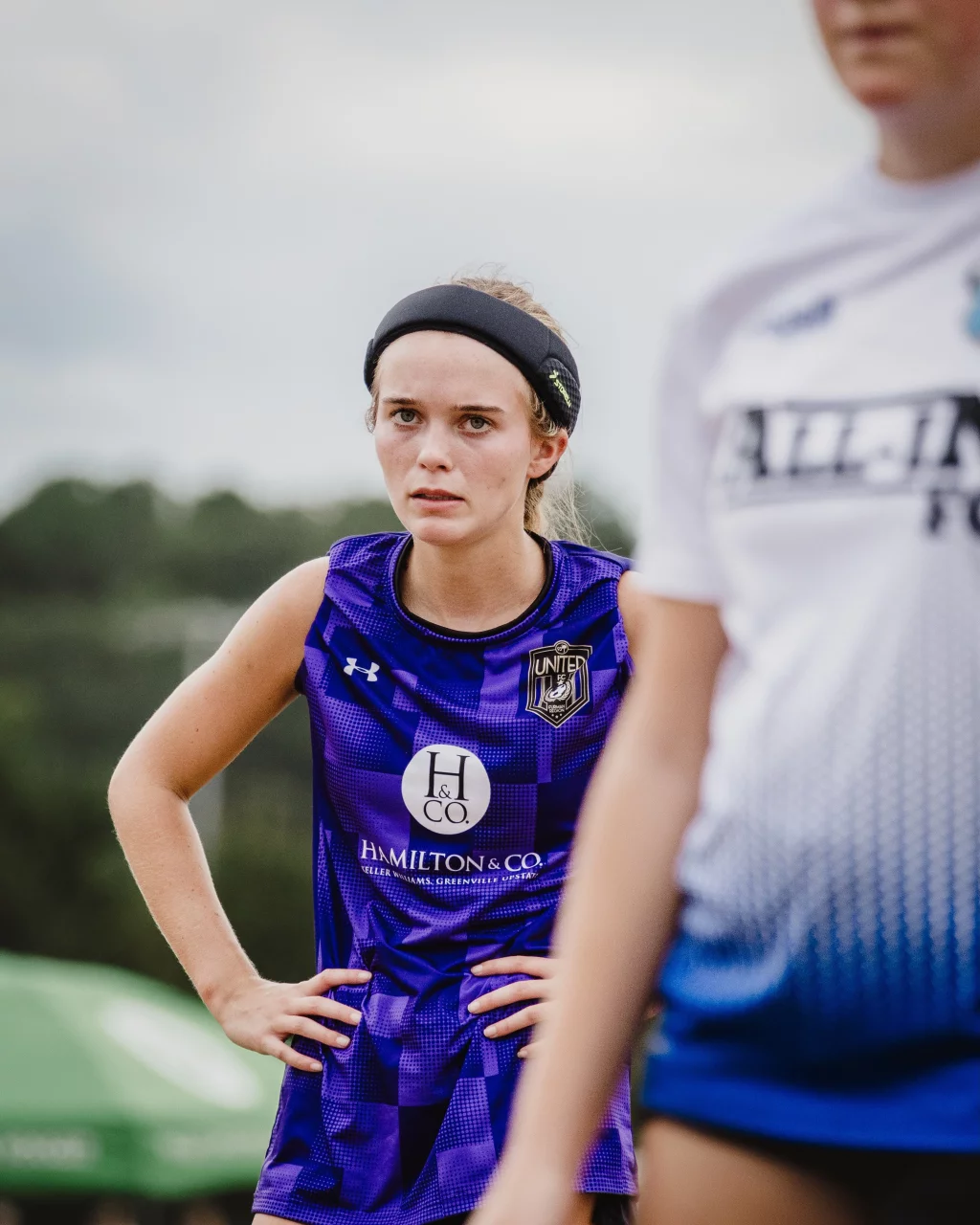 United FC - Furman Region -: A young female soccer player in a purple jersey with hands on hips looks focused, while another player in a white jersey is partially visible in the foreground.