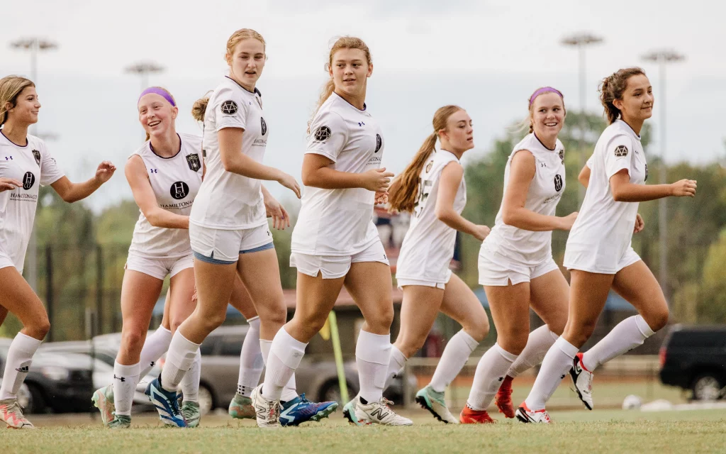 United FC - Furman Region -: A group of girls in white soccer uniforms jogging together on a grass field, smiling and interacting, with trees and light poles in the background.