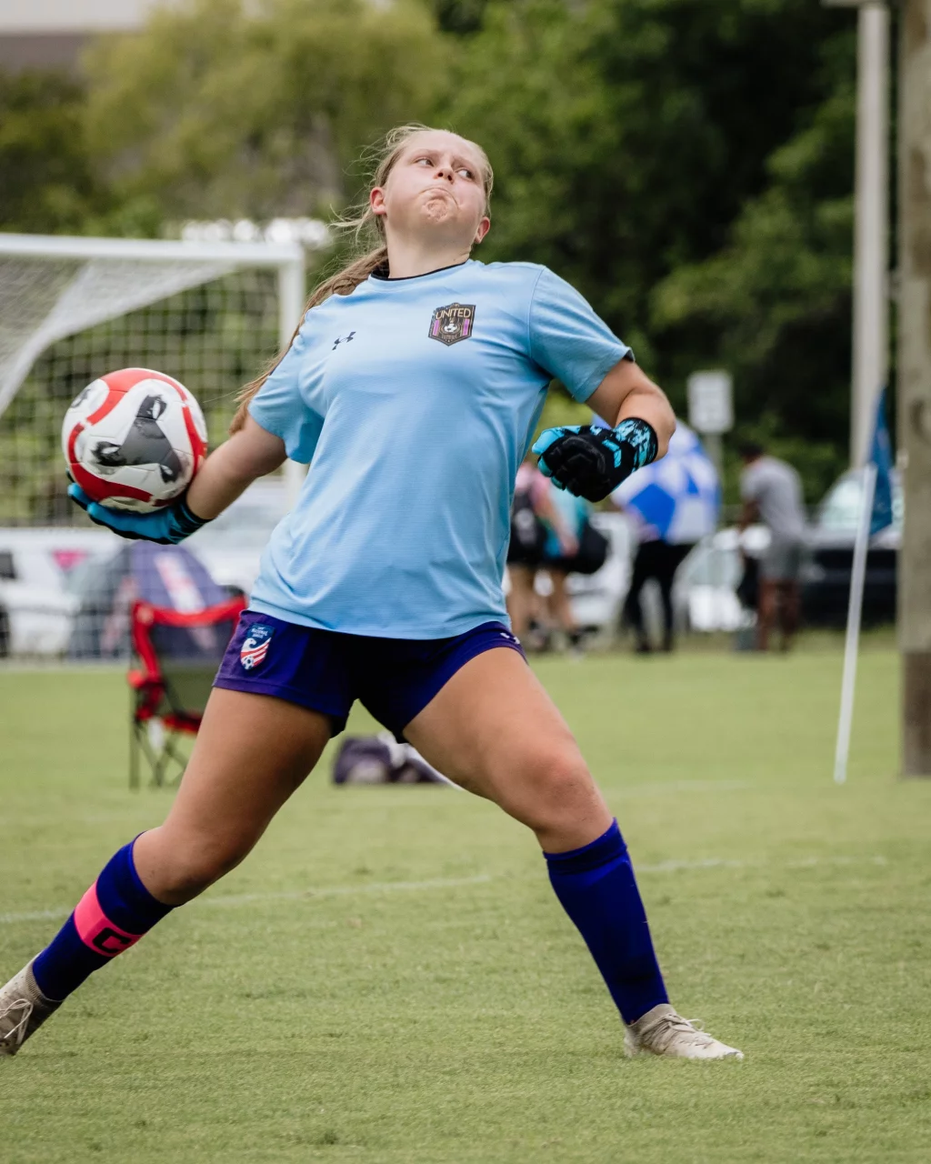 United FC - Furman Region -: A youth soccer goalie in a light blue jersey and gloves is preparing to throw a soccer ball on a grassy field, with a goal and spectators visible in the background.