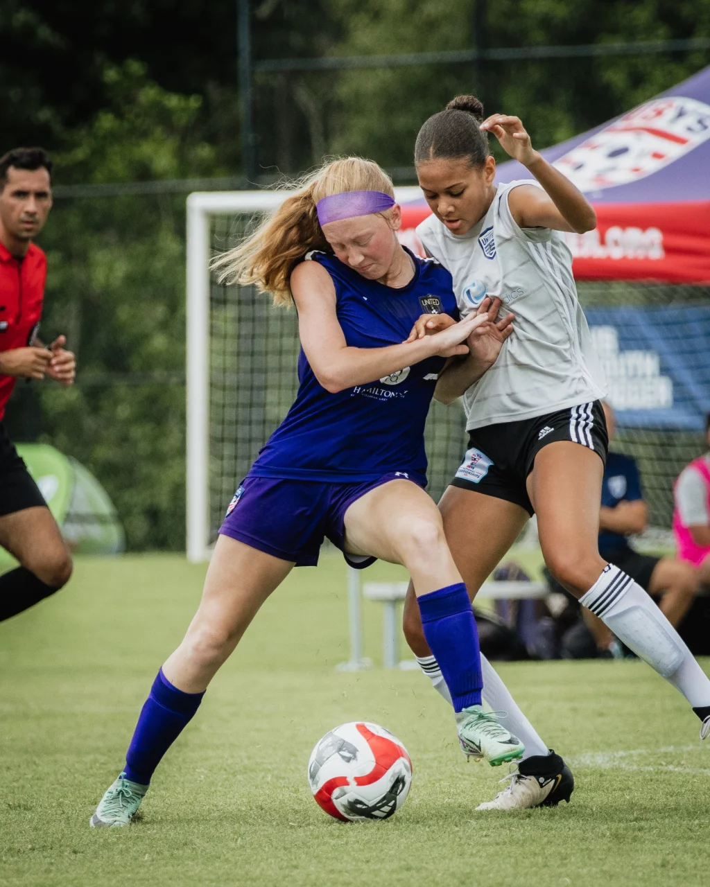 United FC - Furman Region -: Two girls competing for the ball during a soccer match, with one in a blue uniform and the other in a white uniform, both intensely focused and physically engaged. A referee and goalpost are visible in the background.