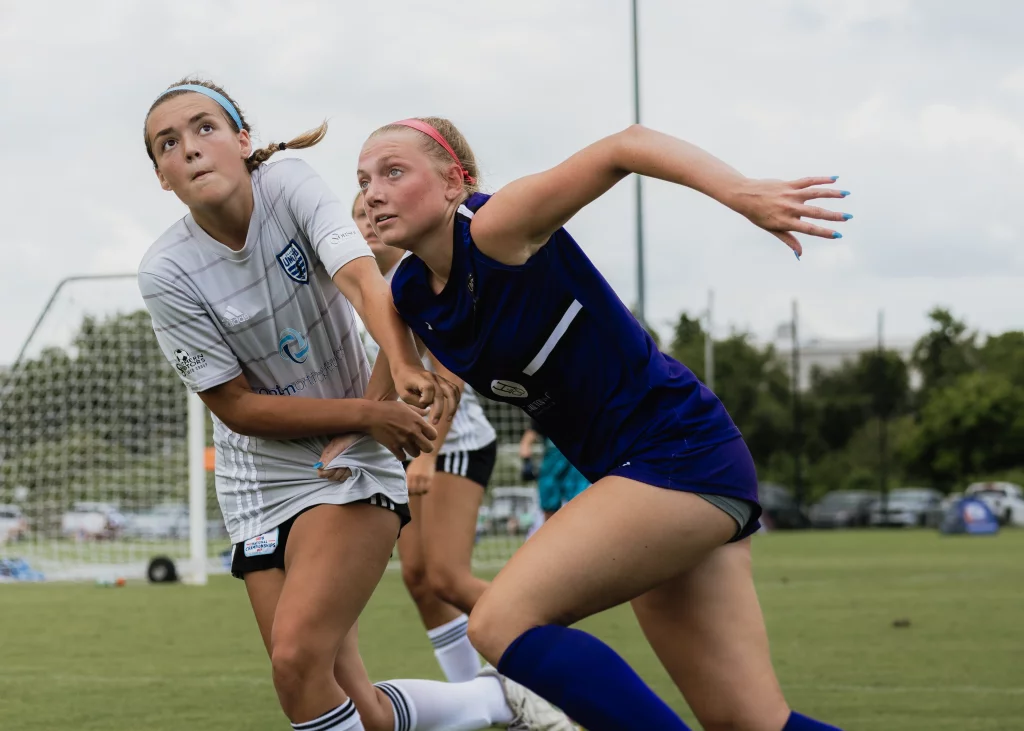 United FC - Furman Region -: Two teenage girls compete for position during a soccer match, one in a white uniform and the other in a blue uniform, with the player in white holding the other&rsquo;s shirt. A goal and players are visible in the background.