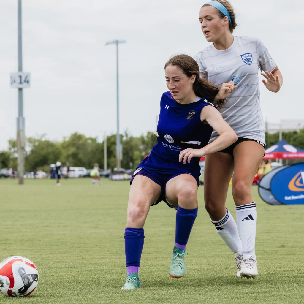 United FC - Furman Region -: Two female soccer players compete for the ball on a grassy field, one in a blue uniform and the other in a white uniform with a headband.