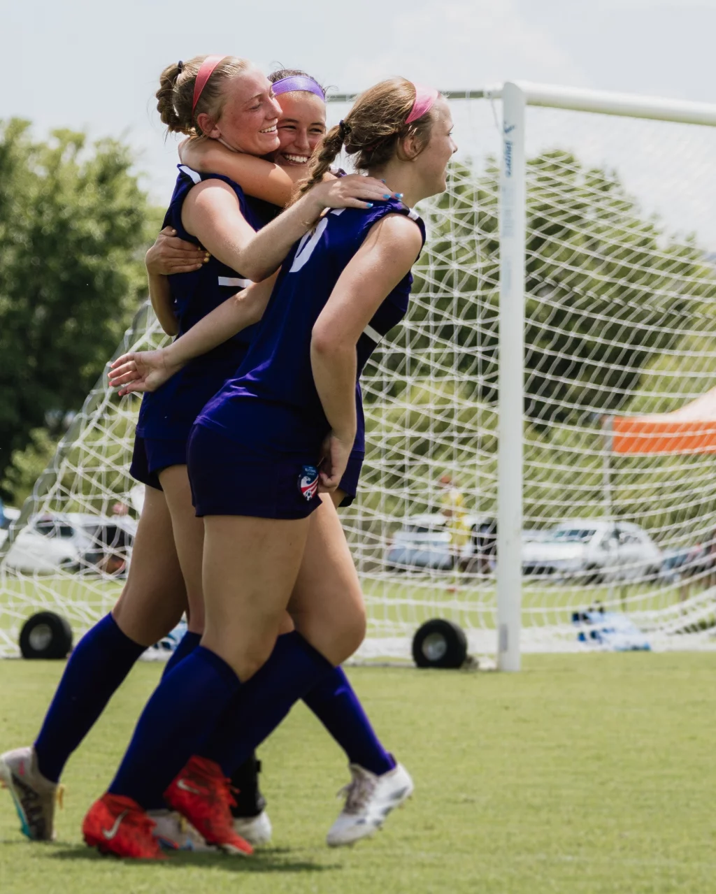United FC - Furman Region -: Three girls in blue soccer uniforms celebrate and hug in front of a goal net on a grassy field.