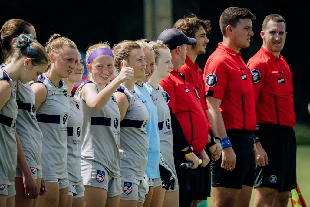 United FC - Furman Region -: A line of female soccer players in gray uniforms stands next to referees in red shirts; one player in the middle gives a thumbs up and smiles.
