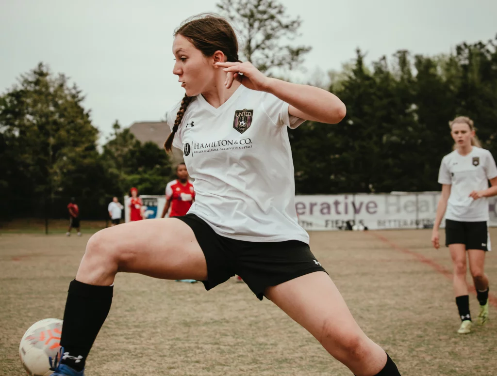 United FC - Furman Region -: A female soccer player in a white jersey and black shorts stretches her leg to control a ball during a game, with teammates and opponents visible in the background on a grassy field.
