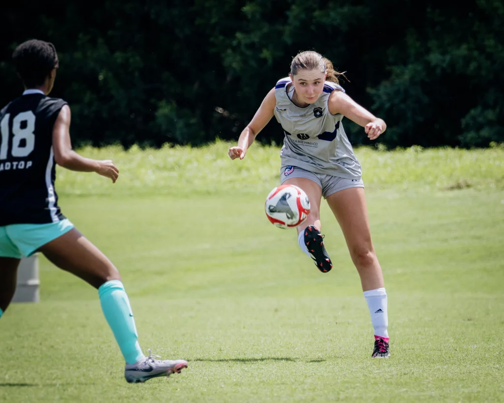 United FC - Furman Region -: Two female soccer players compete on a grassy field; one in a grey uniform is kicking a soccer ball, while the other in a black and turquoise uniform approaches defensively.