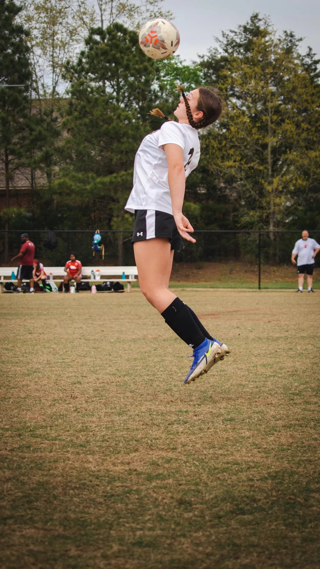 United FC - Furman Region -: Soccer player in mid-air about to head a ball during a game, with spectators and trees in the background.
