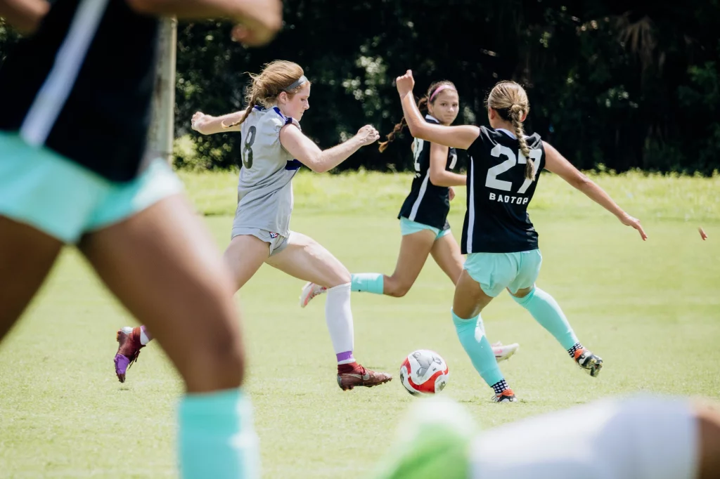 United FC - Furman Region -: Four youth soccer players in action on a grassy field, focused on a red and white soccer ball, with two players in black and aqua uniforms and two in light gray uniforms.