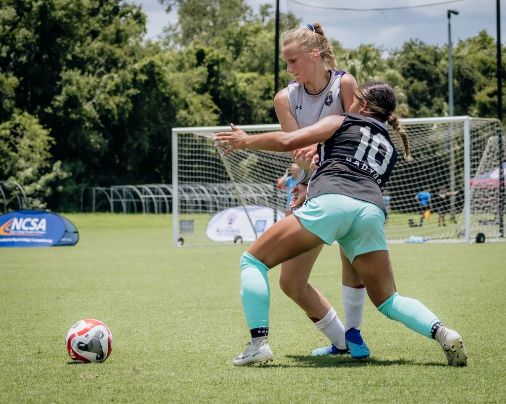 United FC - Furman Region -: Two female soccer players compete for the ball on a grassy field, with one player blocking the other near a goalpost in the background.