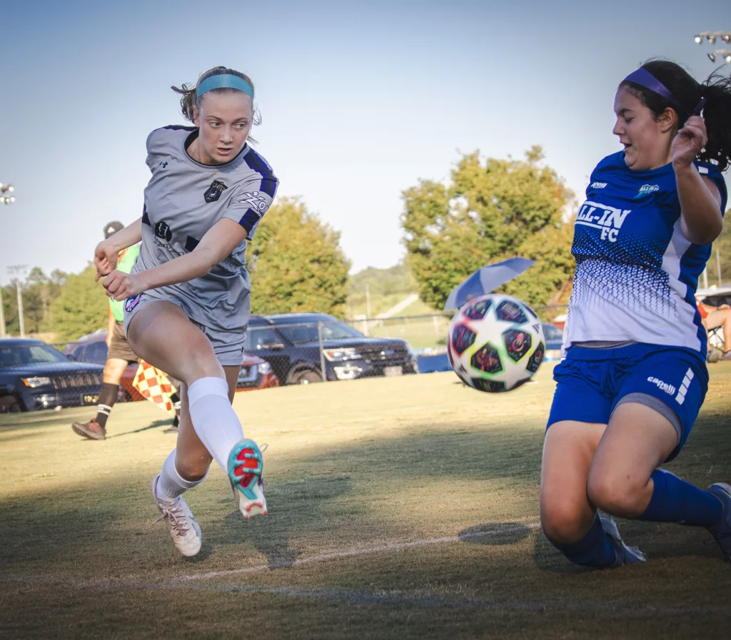 United FC - Furman Region -: Two female soccer players compete for the ball; one in a gray uniform is kicking the ball, while the other in a blue uniform is sliding to block or intercept it on a grassy field, with cars and trees in the background.