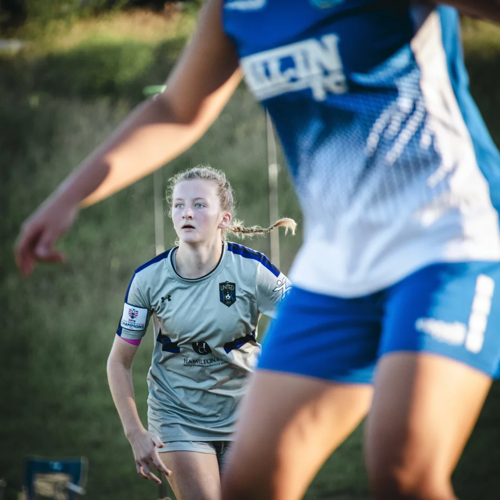 United FC - Furman Region -: A young female soccer player in a gray "United" jersey is focused on the game, with another player in a blue and white uniform partially blocking the foreground. The background is grassy and blurred.