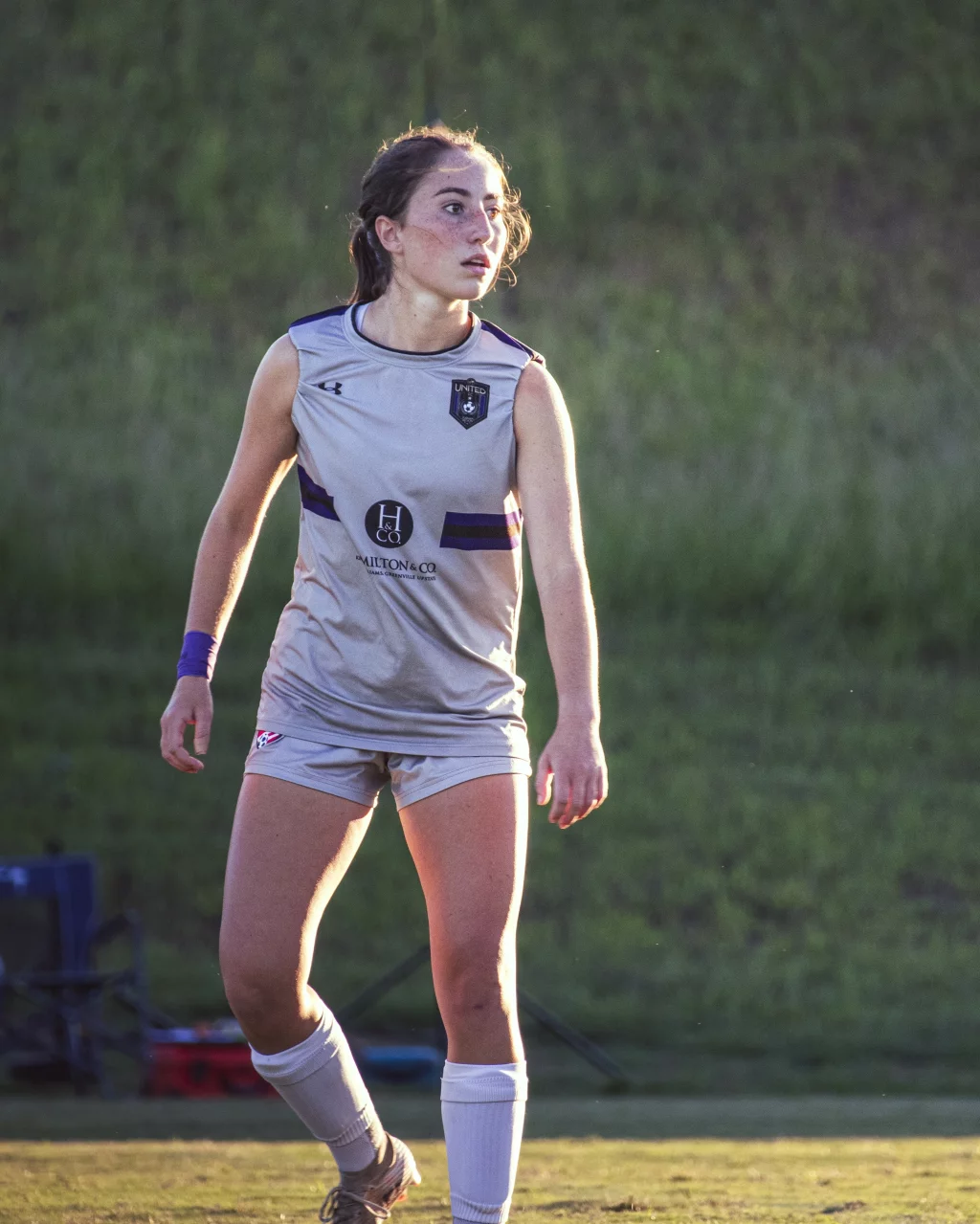 United FC - Furman Region -: A young female soccer player in a gray uniform stands on a grassy field, looking to the side with a focused expression. The background is blurred greenery.