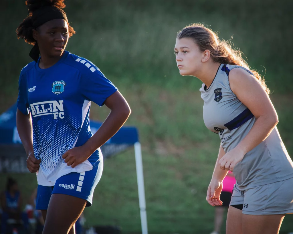 United FC - Furman Region -: Two young female soccer players on a field, one in a blue "All In FC" uniform and the other in a gray "United" uniform, both focused and preparing for action.