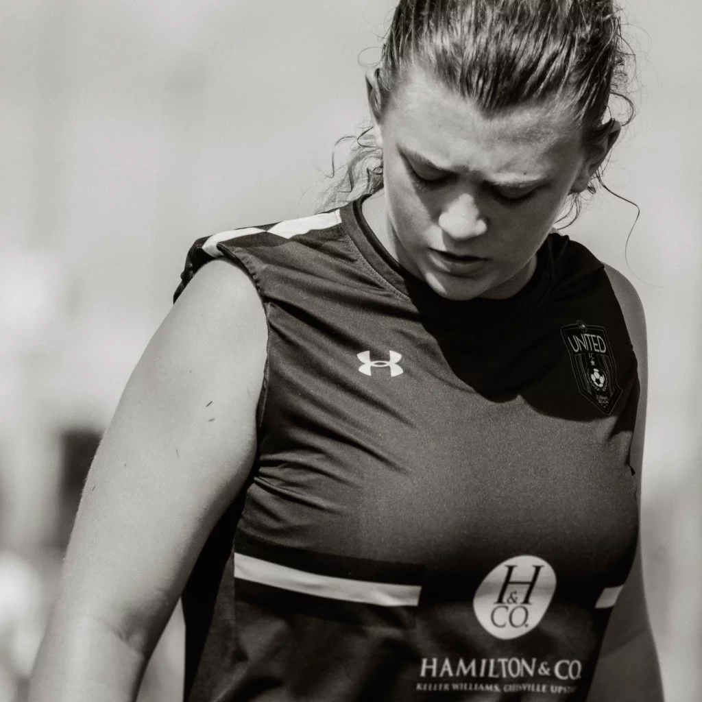 : Black and white photo of a female soccer player looking down, wearing a sleeveless jersey with "UNITED" team crest and sponsorship logos.
