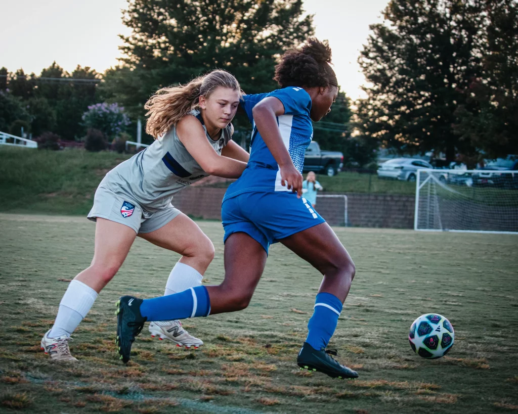 United FC - Furman Region -: Two female soccer players compete for the ball on a grassy field, with one in a gray uniform and the other in blue, as a goal and trees are visible in the background.