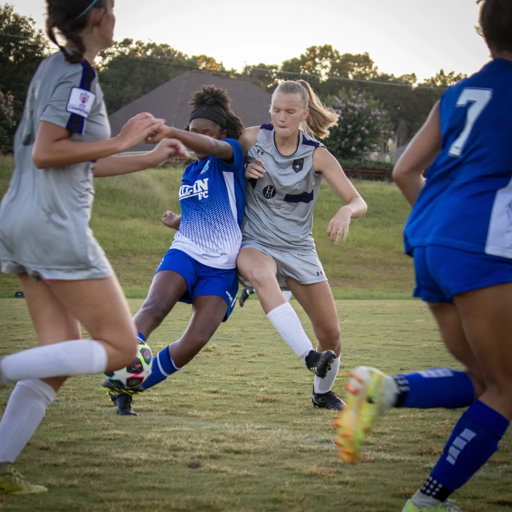 United FC - Furman Region -: Two youth soccer players, one in a blue uniform and one in gray, compete closely for the ball while other players surround them on a grassy field.
