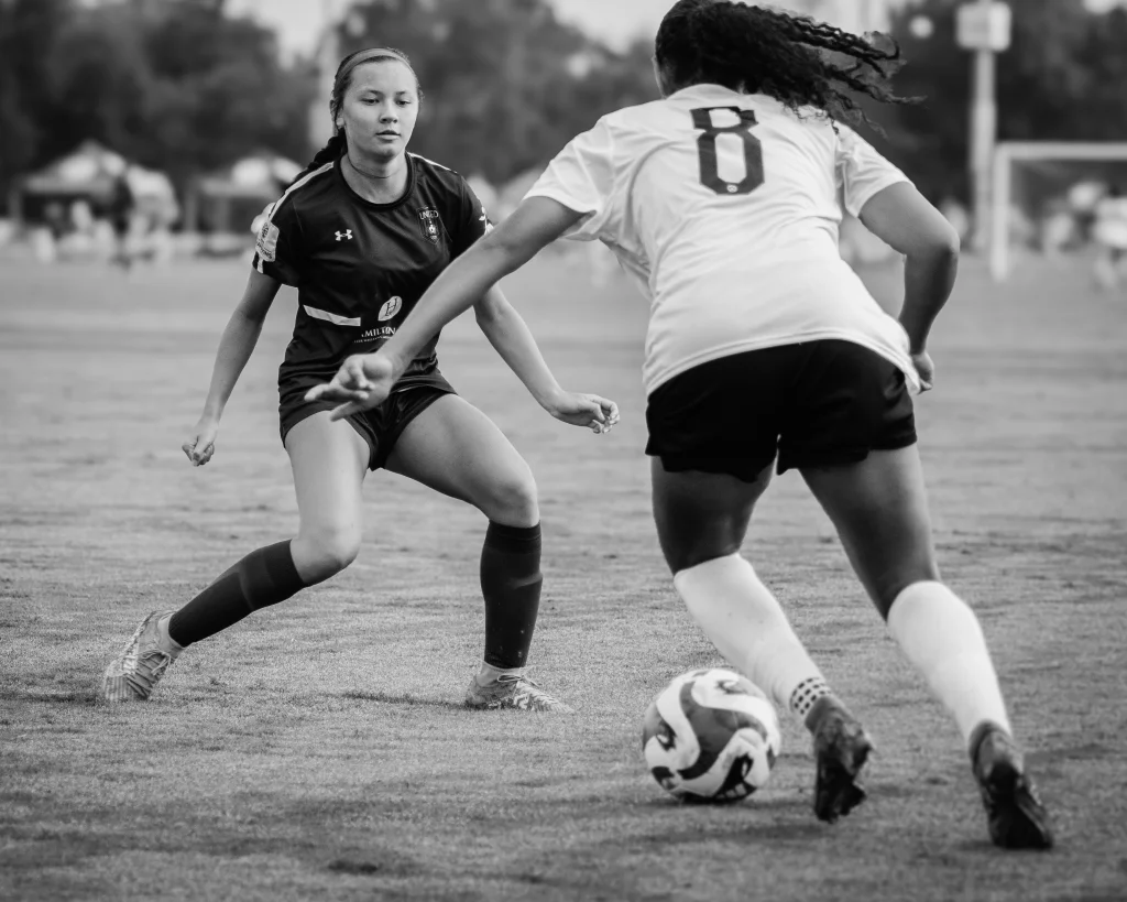 : Two female soccer players face off on a field, one in a dark uniform defending and the other in a light uniform with the number 8 dribbling the ball.