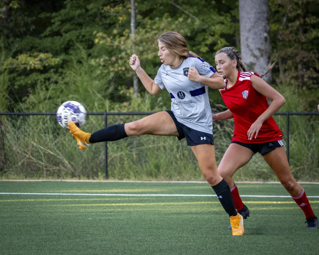 United FC - Furman Region -: Two female soccer players compete for the ball on a field; the player in a gray jersey is mid-kick, while the player in a red jersey is closely defending. Trees and greenery are visible in the background.