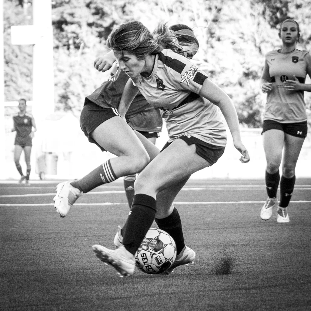 : Black and white photo of two female soccer players in action, closely contesting the ball on a field, with other players visible in the background.