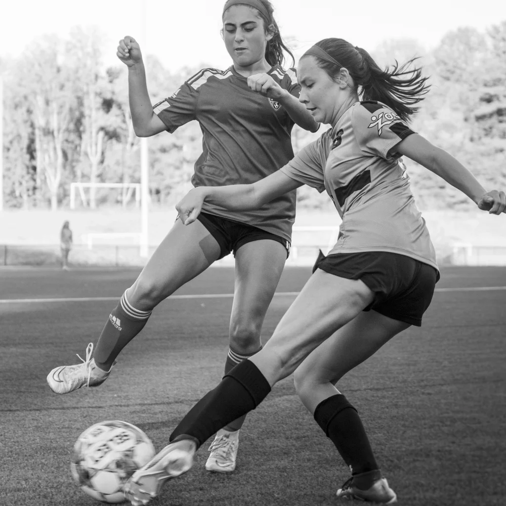: Two teenage girls playing soccer, captured mid-action as they compete for the ball on an outdoor field.