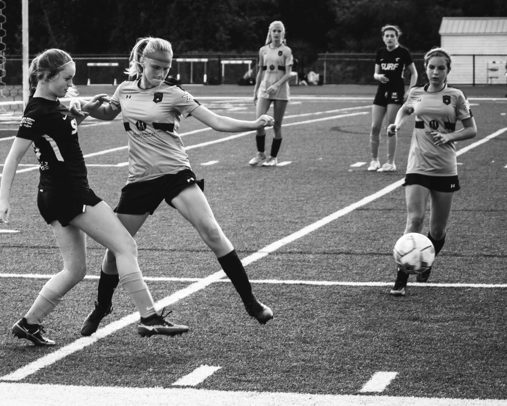 : Black and white photo of a girls' soccer game, showing two players competing for the ball near the sideline while three other players watch in the background.