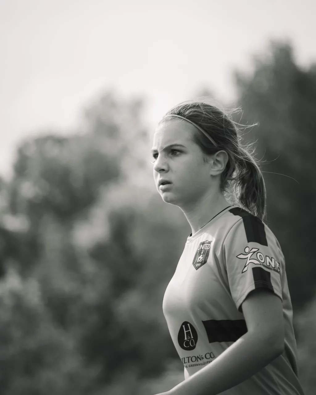 : Black and white photo of a young girl in a soccer jersey, looking focused, with blurred trees in the background.