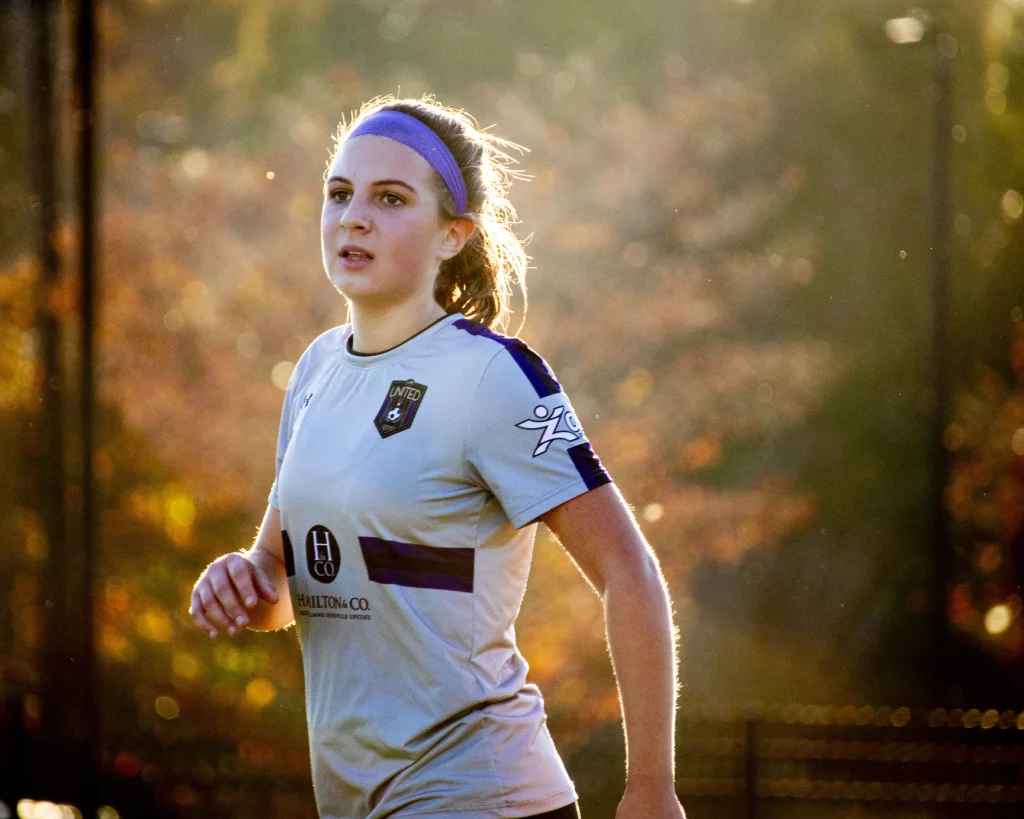 United FC - Furman Region -: A young female soccer player in a light-colored jersey and purple headband is running outdoors, with soft sunlight and autumn trees in the background.