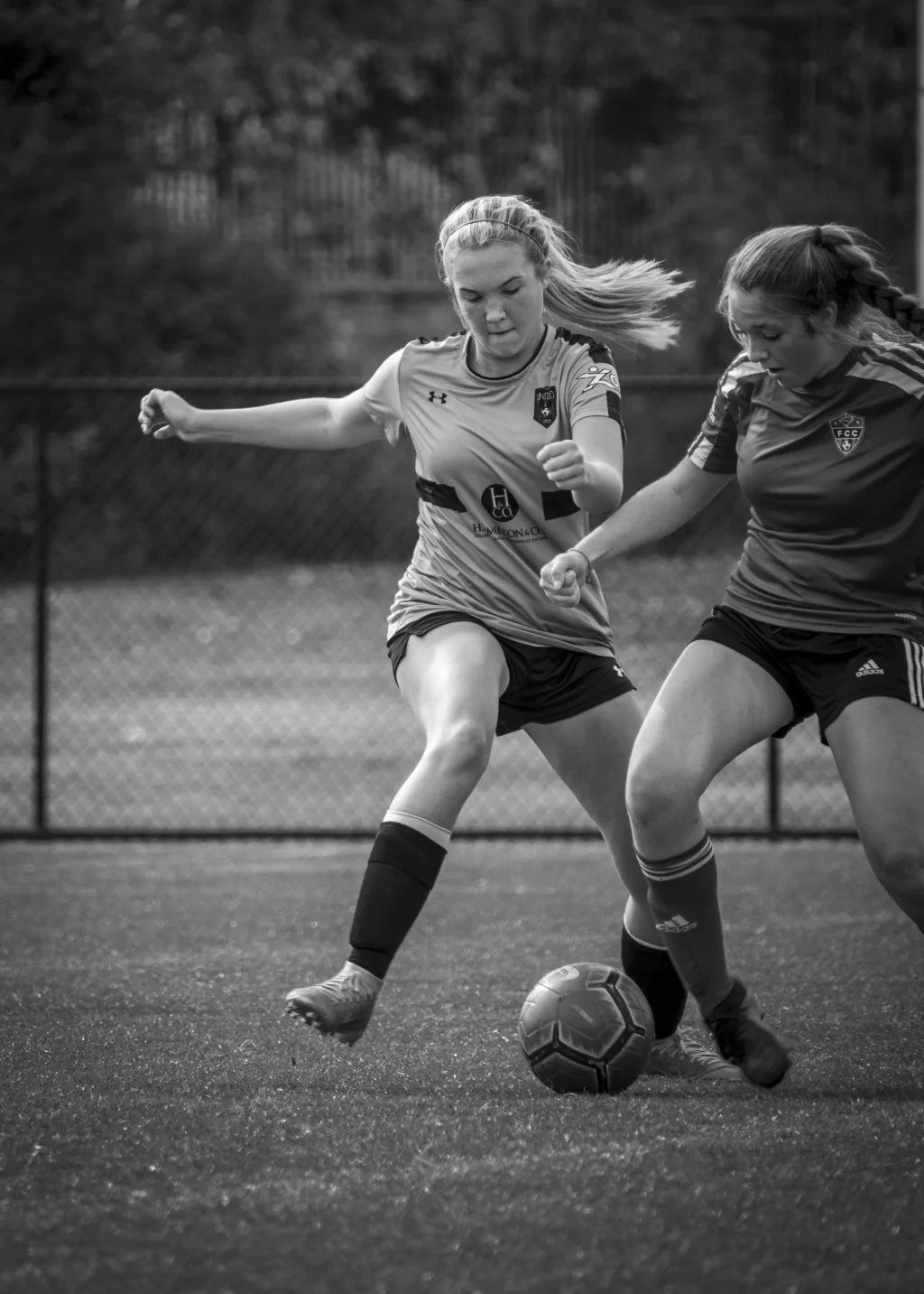 : Black and white photo of two female soccer players in action, both attempting to gain control of the ball on a field.