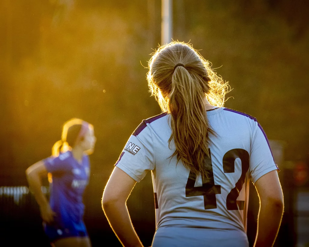 United FC - Furman Region -: Soccer player with long hair in a ponytail, wearing a light-colored jersey with the number 42, stands on a field at sunset with another player blurred in the background.