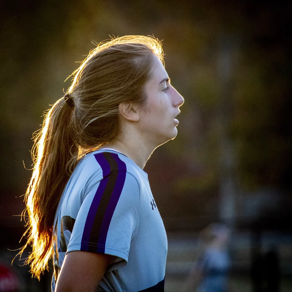 United FC - Furman Region -: A young woman with long hair in a ponytail, wearing a light sports jersey with dark stripes, stands outdoors in profile, illuminated by warm sunlight from behind.